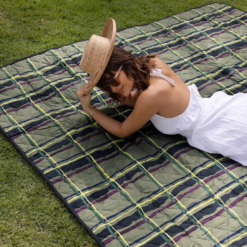 A woman relaxes on a Bambury Felix Picnic Rug, enjoying a sunny day on the grass during an outdoor gathering.