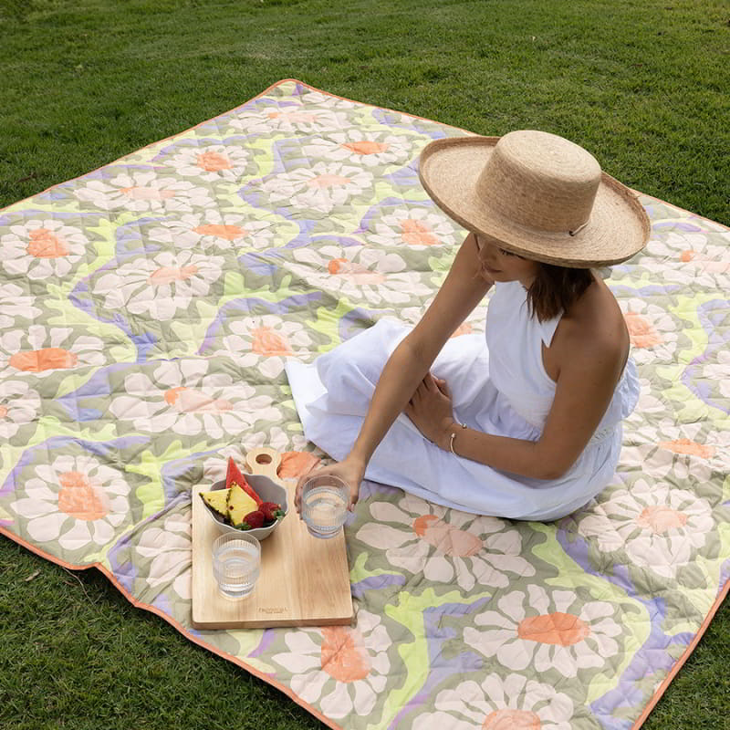  A woman in a hat relaxes on a large, water-resistant Sadie orange picnic rug, enjoying a sunny day outdoors.