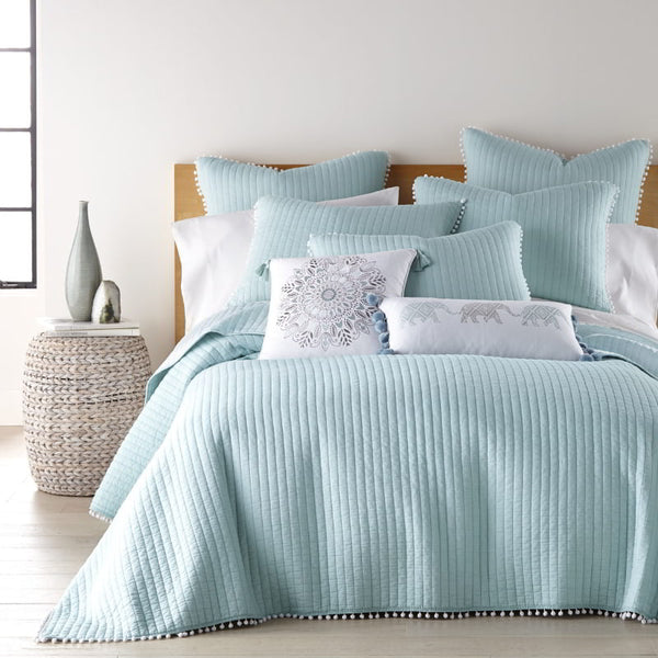 Bright bedroom scene with a light blue comforter and pillows, showcasing playful pom poms for a stylish and comfy vibe.